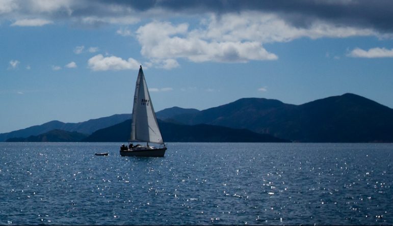 sailboat on blue waters with clouds and dark blue mountains in the background