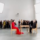 Marina Abramovic, The Artist Is Present, Marina in red sits in one chair, a guest sits in a chair across the table