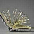 side view of an open laptop with a book on it, pages fanning out from keyboard to screen