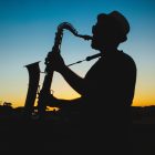 Photograph of a saxophone player silhouetted against a blue and orange sky