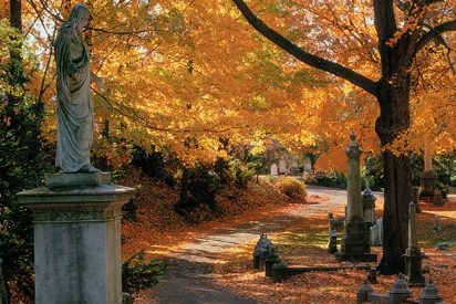 image of a tall stone statue next to a small path in a park at autumn--the surrounding trees are bright orange, covered in autumn foliage