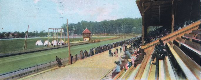 In a vintage postcard style photo, people sit in the bleachers in front of a horseracing track.