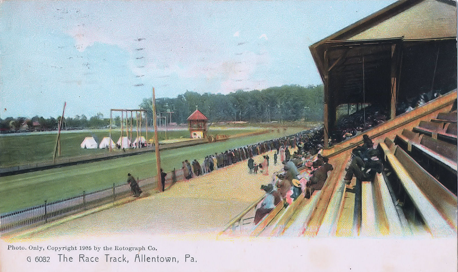 In a vintage postcard style photo, people sit in the bleachers in front of a horseracing track. 