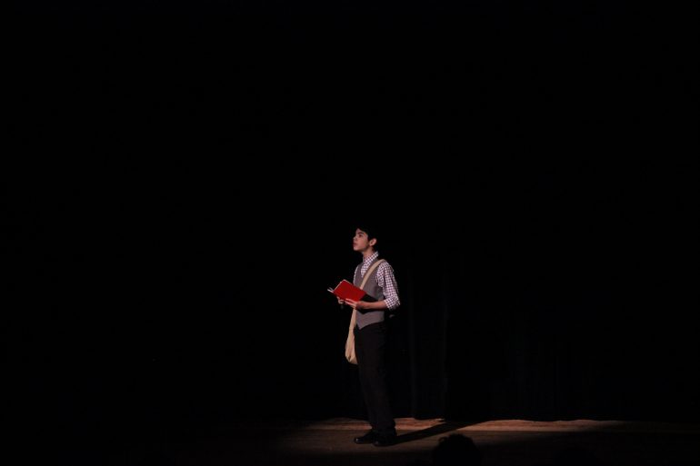 a young person on a stage is in the spotlight, reading from a red book