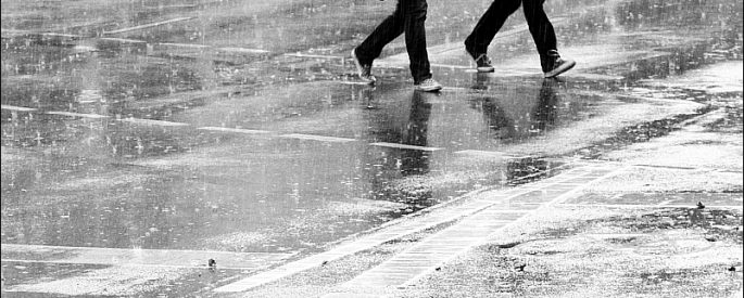 photo of two figures walking in the rain--the photo is in black and white and focused only on their feet and legs as the cross a street
