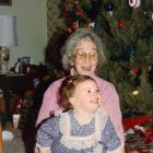 film photo of an older woman in glasses and a pink shirt holding a small child, both in front of a Christmas tree