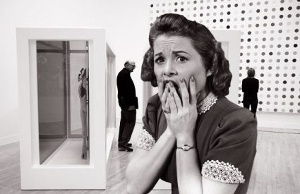 black and white photo of a woman dressed in 50s fashion, with a 50s hairstyle - she appears to be in art museum - a man stands behind her, alongside a wall with a large painting - her expression is that of panic or distress, with her hands covering her mouth