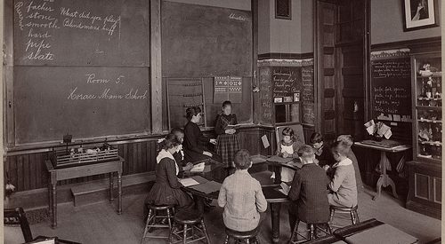 black and white photograph of an old classroom, children sit in a circle with their teacher in front a blackboard with cursive writing across it