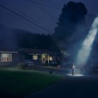 a photograph by the contemporary US photographer Gregory Crewdson of a nighttime suburban scene in which a single figure stands at the end of a driveway with a large beam from some unknown source beaming down on the figure