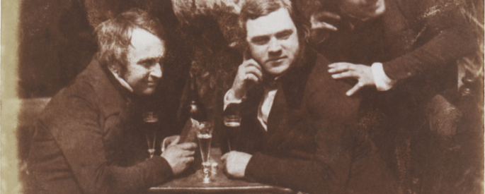 Old, sepia photograph of a group of three men drinking together at a table, one of the men is crouched behind in the other, and is in motion
