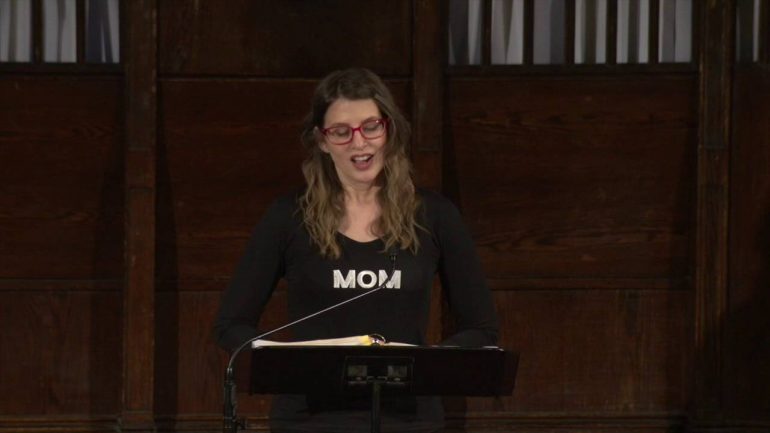 Jackie Mercurio, wearing a shirt that reads "Mom", speaking into a microphone behind a podium 