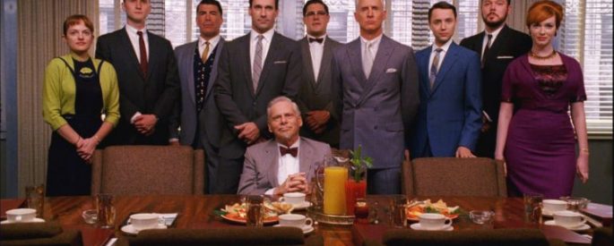 The main characters of Mad Men lined up behind a desk with one man seated at the desk