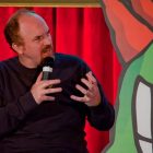 Comedian Louis CK on stage and looking to his left with his hands up