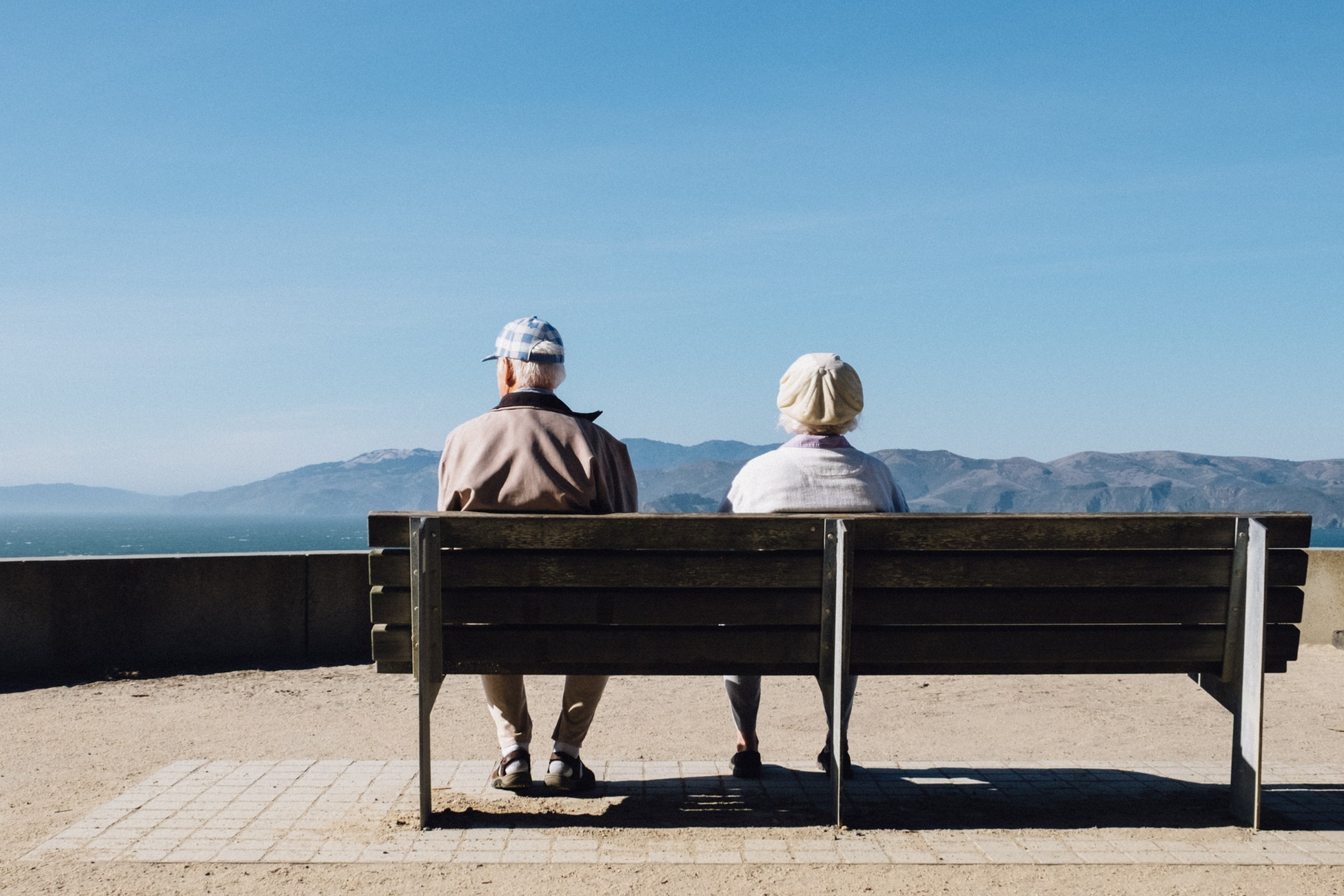 Two older people sitting on a bench with a blue sky above.