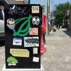 Metal news box on the side of the road covered in stickers, including the logo of The Onion
