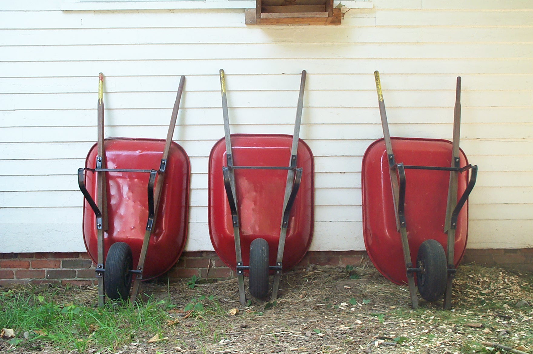 Three red wheelbarrows leaning against the wall of a house.
