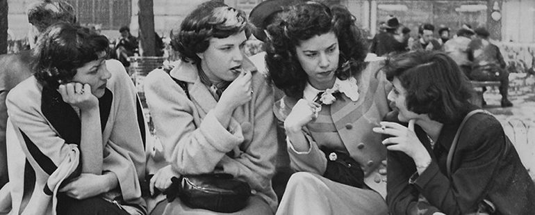 Black and white picture of a group of women sitting on the side of the street chatting with each other.