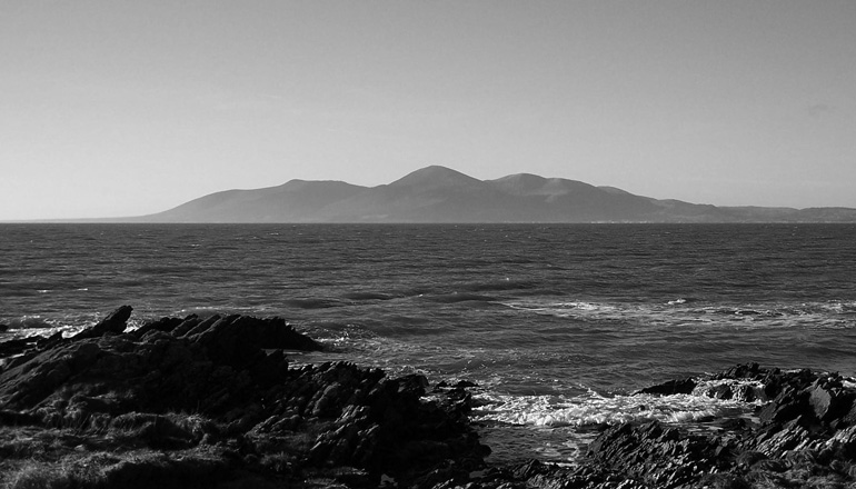Black and white photo of a rocky beach with mountains in the background.
