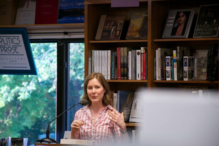 Woman standing at a podium in front of a bookshelf.