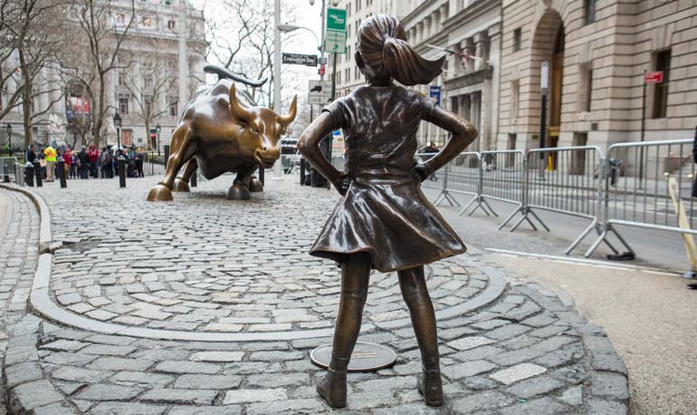 Statue of a young girl standing in front of another statue of a charging bull.