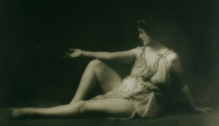 Woman wearing a dress and extending a hand while looking away from the camera.
