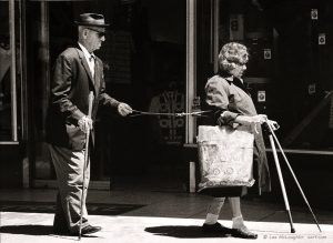 Old man in a suit holding on to a harness attached to an old woman holding walking canes.
