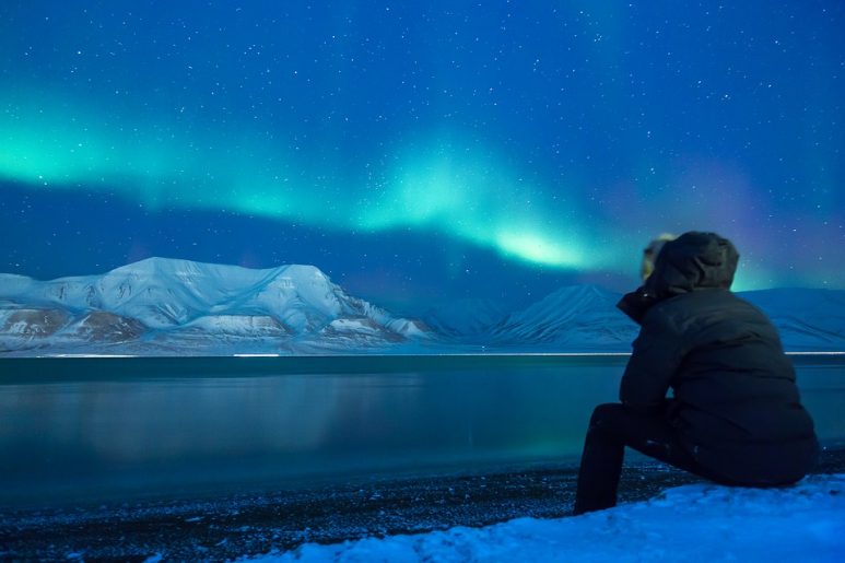 Person sitting on snow covered ground watching the Northern Lights.