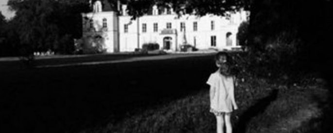 in black and white, a young girl, back to camera, looks at a white mansion-like building some distance away