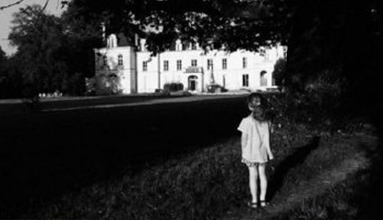 in black and white, a young girl, back to camera, looks at a white mansion-like building some distance away