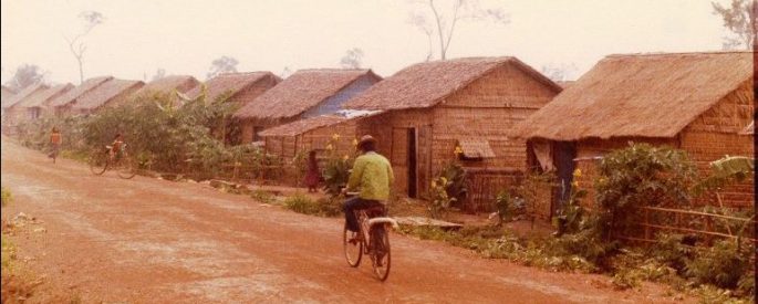 person rides bike on dirt road lined with houses