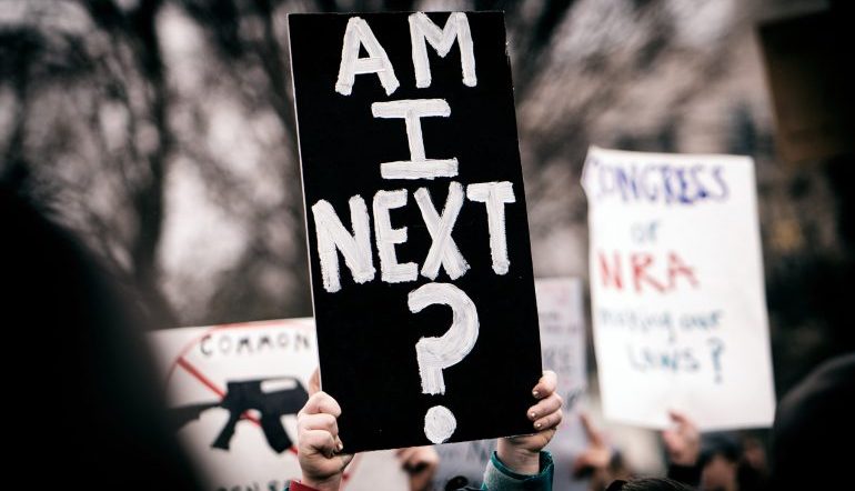 Hands holding a black sign that says "Am I next?" in white lettering