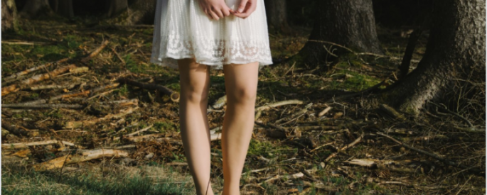 person in a white dress standing in a forest; we see only their legs, hands, and the forest floor
