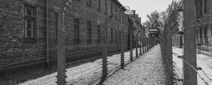 Black and white photograph of a pathway in Auschwitz with barbed wire fencing on both sides.