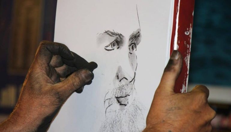 A hand sketching a charcoal portrait of a face