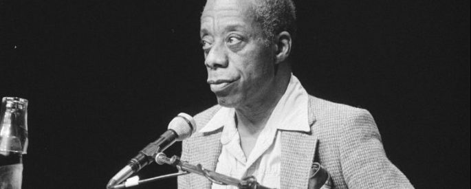Black and white photo of James Baldwin standing behind a podium speaking into a microphone