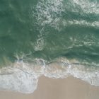 an aerial photograph of a green-ish ocean wave coming in to sand