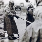 historical black and white photograph of a a figure in a hazmat suit spraying the wheels of a car out of frame with a hose - other people in the photograph are also wearing hazmat suits, the images comes from a collections of the Chernobyl accident from the Ukrainian Society for Friendship and Cultural Relations with Foreign Countries