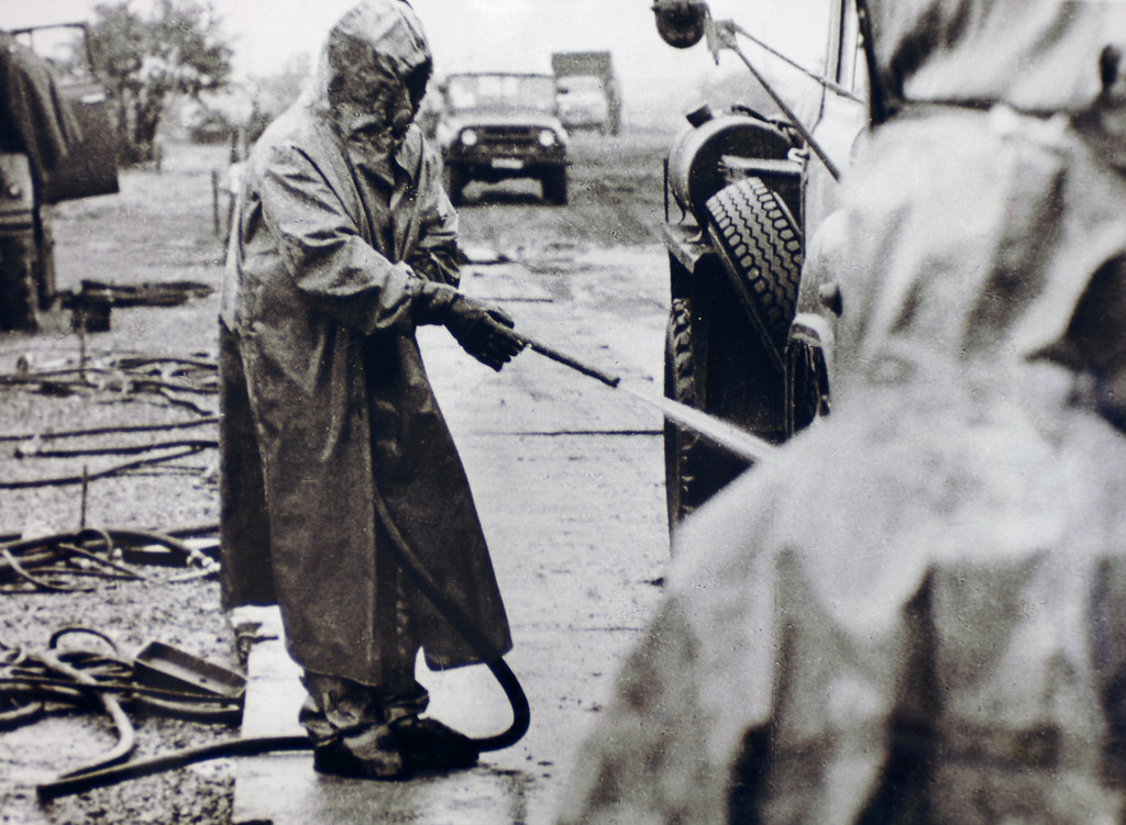 historical black and white photograph of a a figure in a hazmat suit spraying the wheels of a car out of frame with a hose - other people in the photograph are also wearing hazmat suits, the images comes from a collections of the Chernobyl accident from the Ukrainian Society for Friendship and Cultural Relations with Foreign Countries