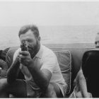 black and white photo of Hemingway pointing a gun at the taker of the photograph, the gun covering half of his face - he sits aboard a boat, accompanied by a male friend