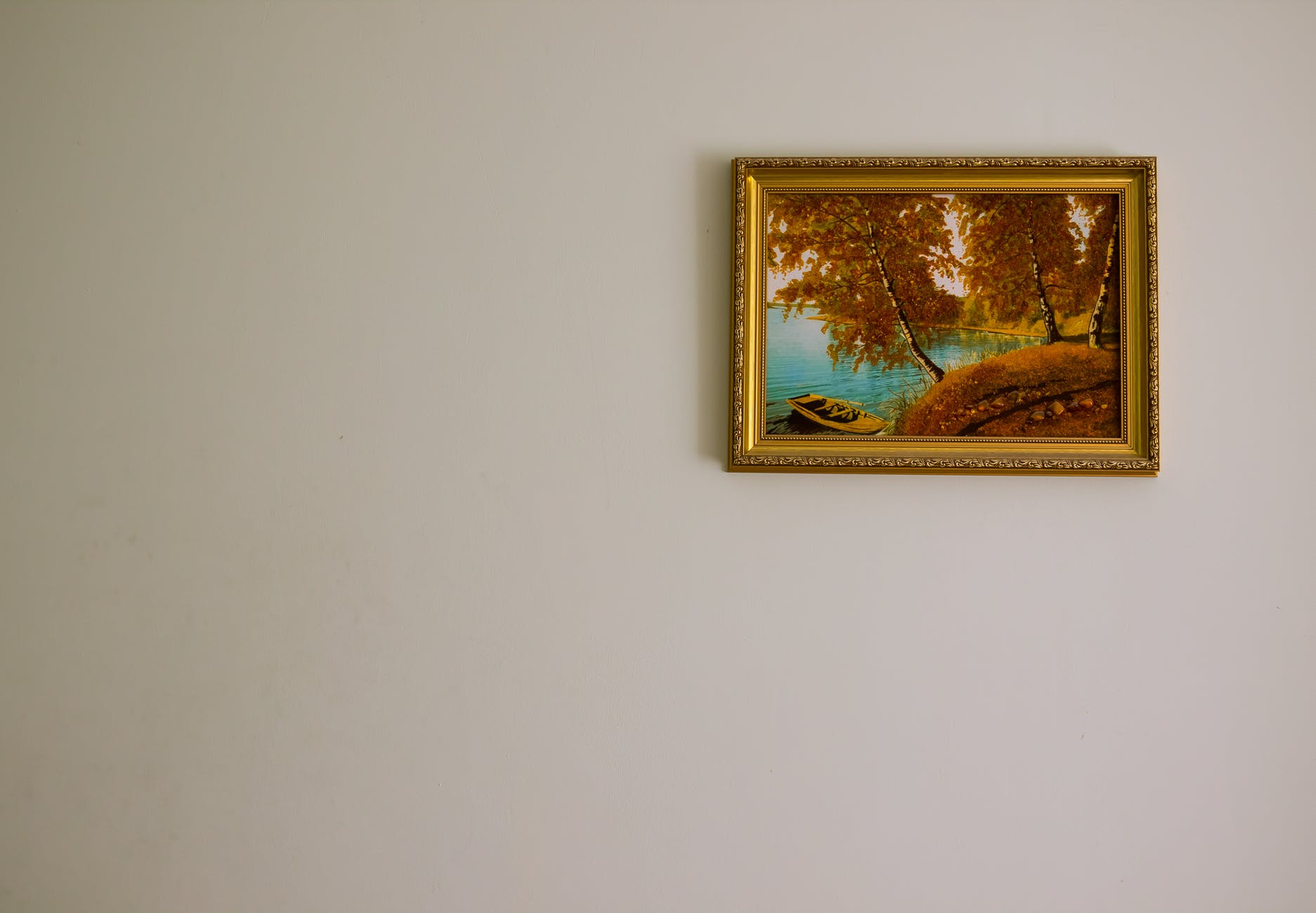 a single painting of a boat on a lake shaded by autumn trees in a gold frame hangs on a bare, beige wall 