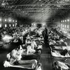 a black and white photograph of a makeshift hospital interior--rows and rows of hospital beds contain some patients who are sitting up in their beds and nurses in old, white nurse uniforms weaving throughout the rows of beds
