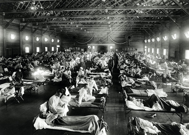 a black and white photograph of a makeshift hospital interior--rows and rows of hospital beds contain some patients who are sitting up in their beds and nurses in old, white nurse uniforms weaving throughout the rows of beds