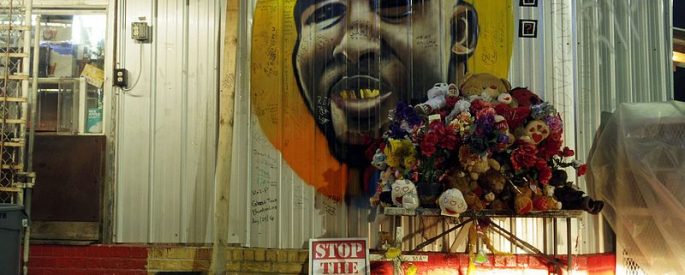 a mural of Alton Sterling on the white-gray walls of a building; beneath is a stand with a pile of bouquets of flowers and stuffed animals, a pair of headlights shine off the base of the wall next to a sign that, in white and red lettering reads, "STOP THE VIOLENCE" with an illustration of a gun