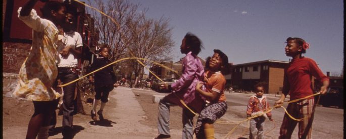 on a sidewalk, a group of young Black girls play double Dutch