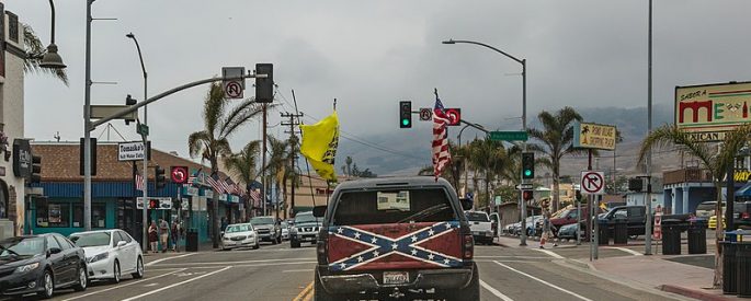 photograph of a pick up truck with a confederate flag on the tailgate. the truck is driving own a street--the background consists of an intersection and palm trees