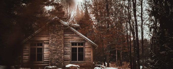 photograph of a cabin in snowy woods, the photo seems to be taken through the trees