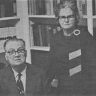 a black and white photograph of an older man and woman in front of a bookshelf
